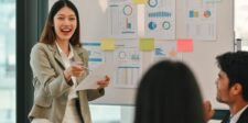 Woman presenting in an office meeting while holding a paper, with charts and graphs pinned on a whiteboard behind her