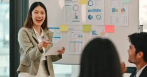 Woman presenting in an office meeting while holding a paper, with charts and graphs pinned on a whiteboard behind her