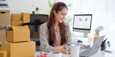 Small business owner working on a laptop beside shipping boxes, with business analytics visible on a desktop monitor in the background.