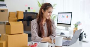 Small business owner working on a laptop beside shipping boxes, with business analytics visible on a desktop monitor in the background.