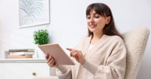 Woman smiling while using a tablet in a bright home office, suggesting someone reading or writing thank you letters online.
