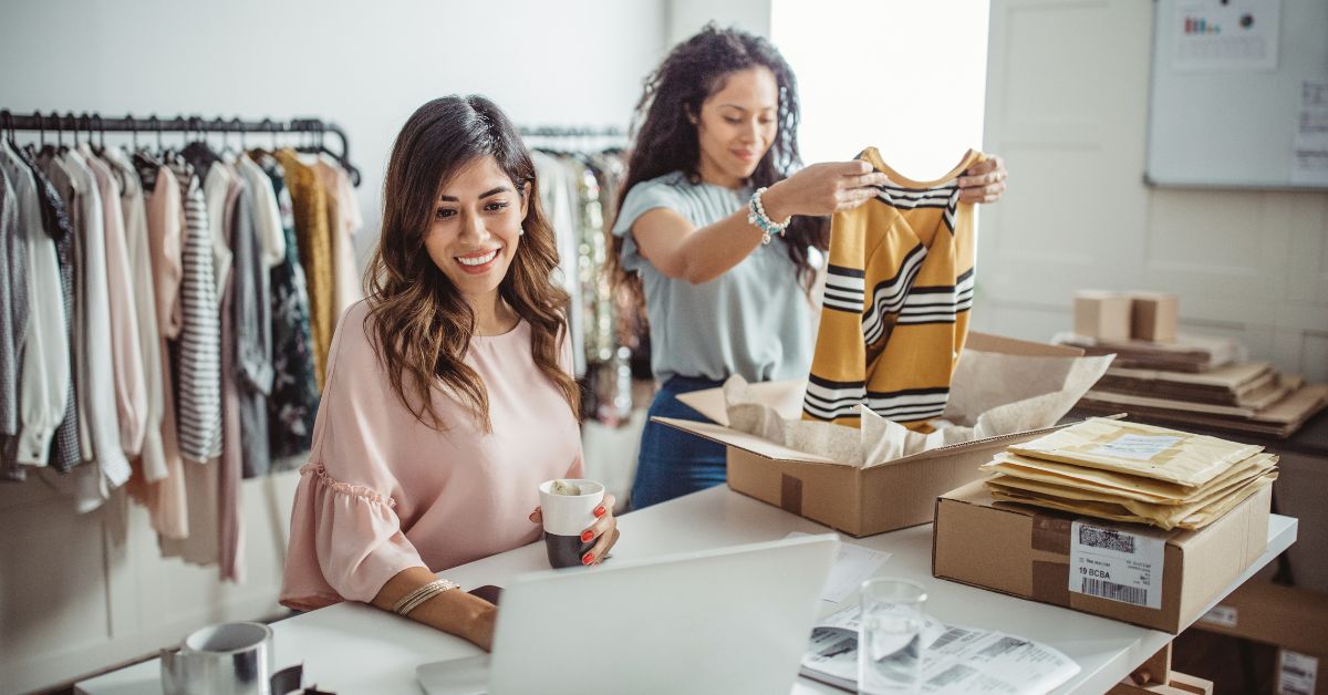 Small business owners preparing clothing orders at a worktable with shipping boxes and apparel racks in the background.