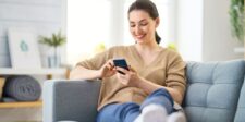 AI Search Isn't a Trend Anymore — It's How People Find You Now Woman sitting on a couch and smiling while using a smartphone, representing how people now use AI and mobile tools to search for information.