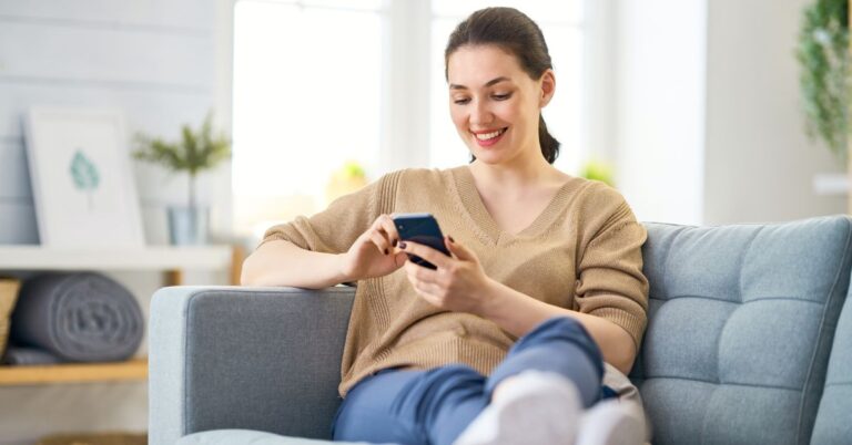 Woman sitting on a couch and smiling while using a smartphone, representing how people now use AI and mobile tools to search for information.
