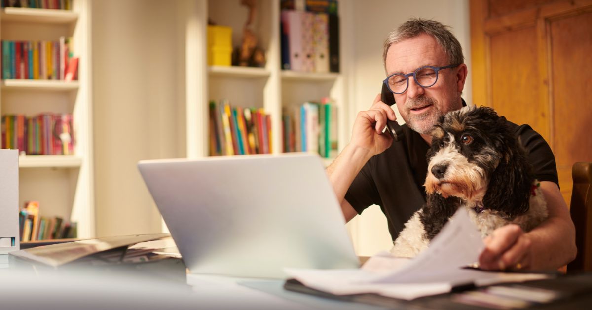 Man working on a laptop while talking on the phone, with a small dog sitting beside him at a desk.