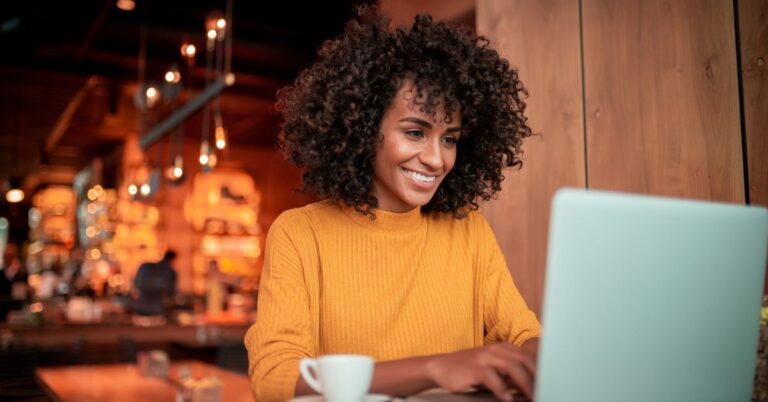 Woman in a yellow sweater smiling while using a laptop in a café