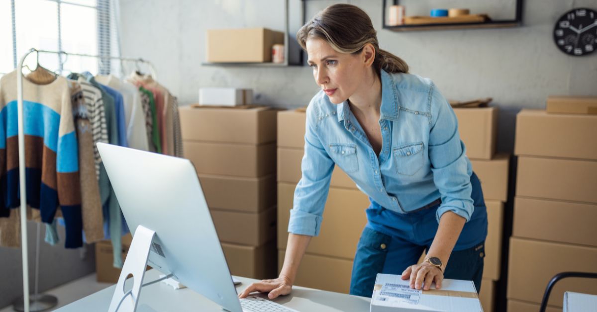 Small business owner working at a desktop computer beside shipping boxes and clothing inventory, representing online business management.
