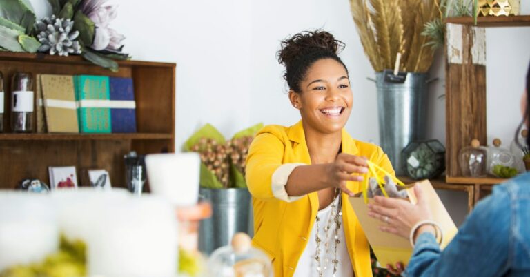 Smiling retail worker handing a gift bag to a customer inside a bright boutique shop.