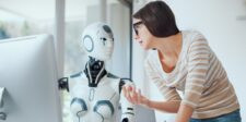 Woman speaking with a humanoid robot beside a desktop computer in a bright office setting.