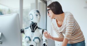 Woman speaking with a humanoid robot beside a desktop computer in a bright office setting.