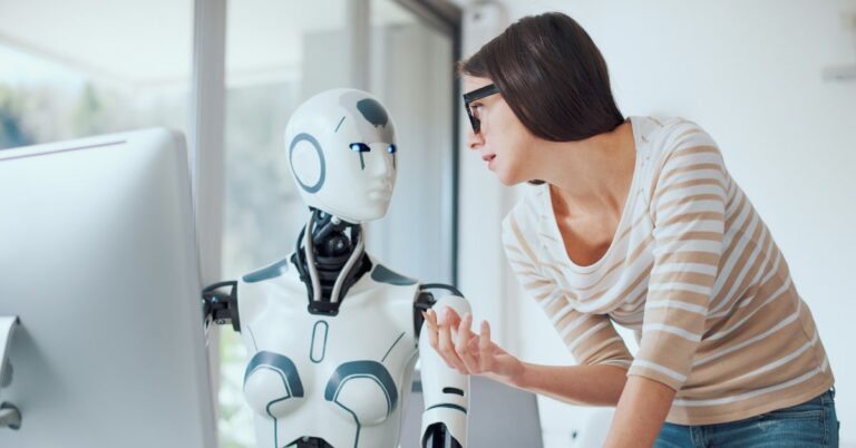 Woman speaking with a humanoid robot beside a desktop computer in a bright office setting.