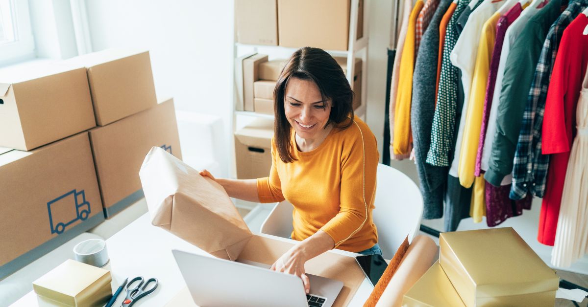 Woman packing an online clothing order in a small business workspace with shipping boxes, clothes, and a laptop.