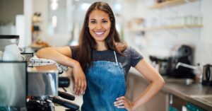 Smiling café worker in an apron standing beside an espresso machine in a bright coffee shop.