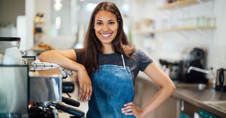 Smiling café worker in an apron standing beside an espresso machine in a bright coffee shop.