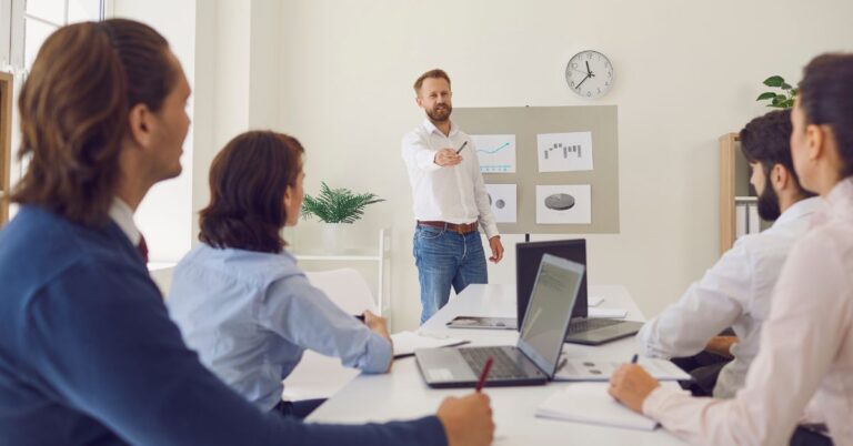 Business professional presenting charts and graphs to a team seated around a conference table with laptops in a modern office setting