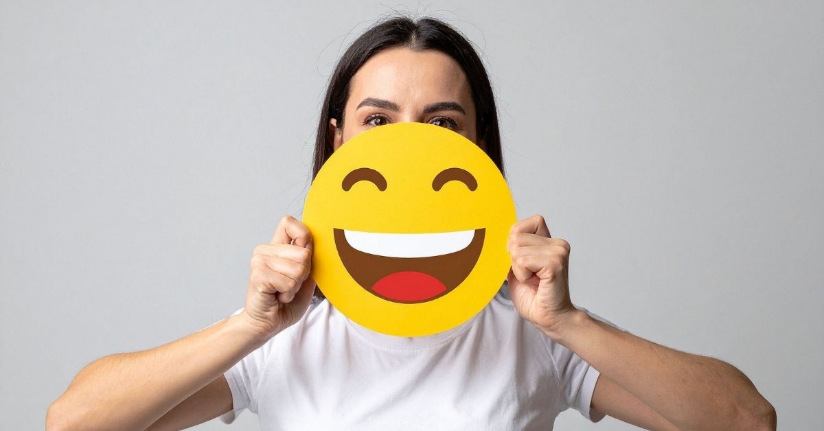 Person holding a large smiling emoji sign in front of their face against a plain gray background.