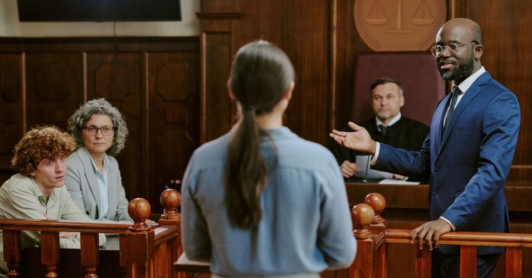 Courtroom scene showing a lawyer speaking before a judge and other people during a legal proceeding.
