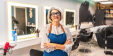 Smiling woman wearing glasses and a blue apron standing with her arms crossed inside a bright, modern hair salon.
