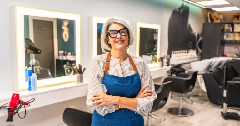 Smiling woman wearing glasses and a blue apron standing with her arms crossed inside a bright, modern hair salon.