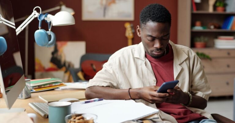 Young man sitting at a desk and looking at his phone in a creative home office.