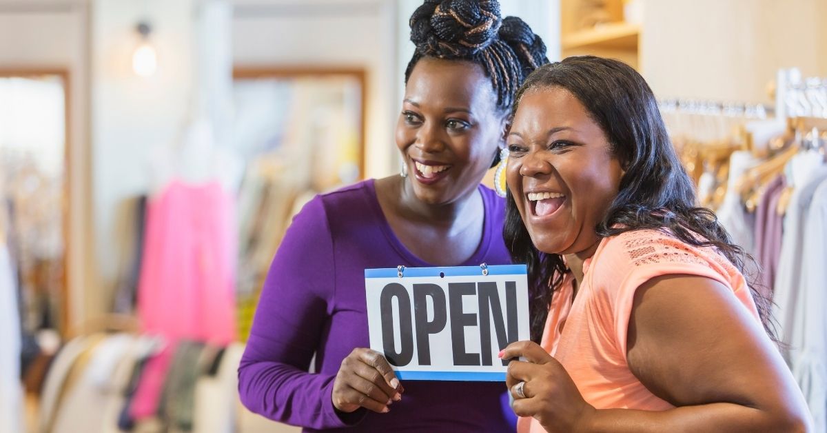 Two smiling women holding an “Open” sign inside a clothing shop.