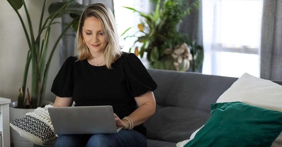 Woman sitting on a couch and working on a laptop in a bright home office setting.