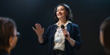 The money skills nobody calls money skills Woman speaking into a microphone during a presentation, representing communication and public speaking as practical money skills.