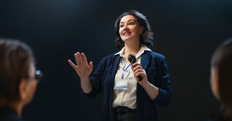 Woman speaking into a microphone during a presentation, representing communication and public speaking as practical money skills.
