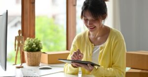 Woman using a tablet at a home office desk with moving boxes nearby, representing someone starting or managing a one-person business.