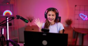 A smiling video creator wearing headphones waves at the camera while recording in a colorful home studio.
