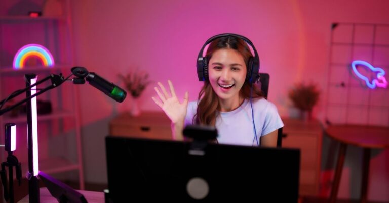 A smiling video creator wearing headphones waves at the camera while recording in a colorful home studio.