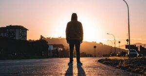 Person in a hooded sweatshirt standing alone in the middle of a road at sunset.