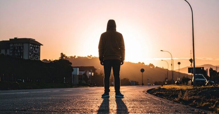 Person in a hooded sweatshirt standing alone in the middle of a road at sunset.