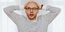 Surprised man wearing glasses with both hands on his head against a bright white background.