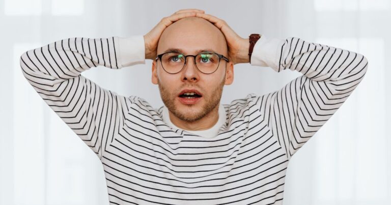 Surprised man wearing glasses with both hands on his head against a bright white background.