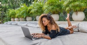 Woman lying on an outdoor lounge chair while working on a laptop.