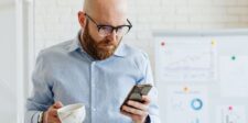 The Google Ads setting that could double your cost per customer overnight Man looking at his phone while holding a coffee cup, with charts in the background, representing a business owner or marketer reviewing advertising performance.