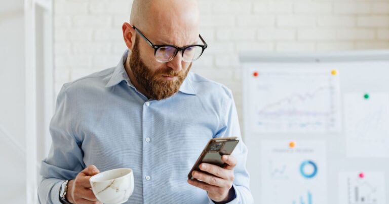 Man looking at his phone while holding a coffee cup, with charts in the background, representing a business owner or marketer reviewing advertising performance.
