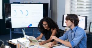 Two people reviewing marketing analytics on a large screen in an office.