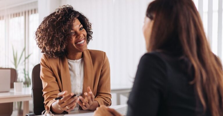 Two women having a conversation across a desk in a bright office.