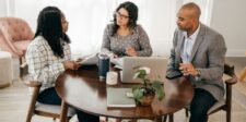 How much do marketing agencies charge? Three people sitting around a table with a laptop and documents during a business meeting.