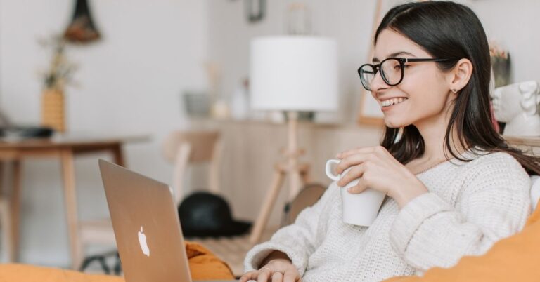 Smiling woman wearing glasses and holding a mug while using a laptop at home.