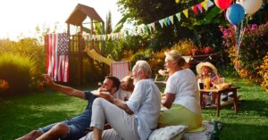 Family and friends gathered outside for a sunny backyard celebration with food, balloons, flags, and festive decorations.