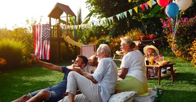 Family and friends gathered outside for a sunny backyard celebration with food, balloons, flags, and festive decorations.