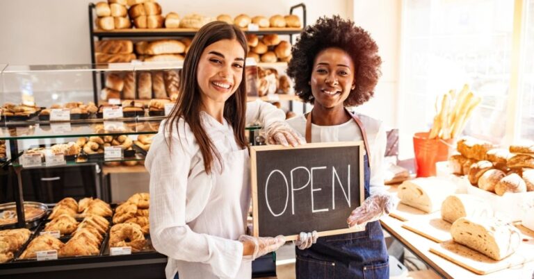 Two bakery workers smiling while holding an open sign in front of a pastry display.