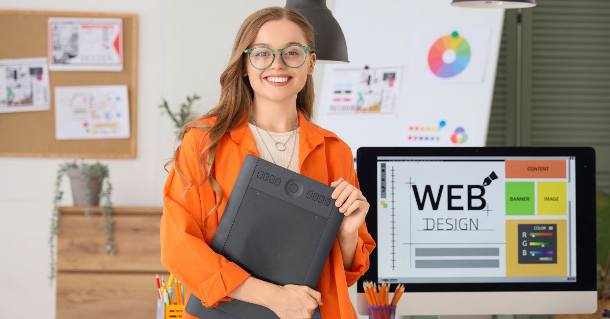 Smiling web designer holding a drawing tablet beside a desktop monitor showing a web design layout.