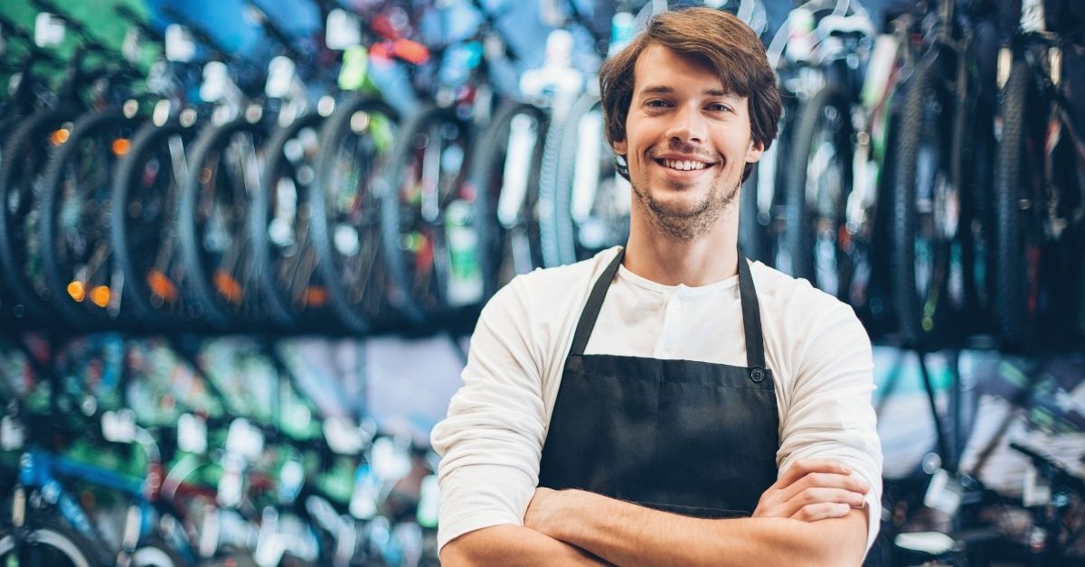 Smiling man in a black apron standing with his arms crossed inside a bicycle shop.