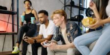 Group of friends playing video games together in a living room, with controllers, drinks, and snacks.