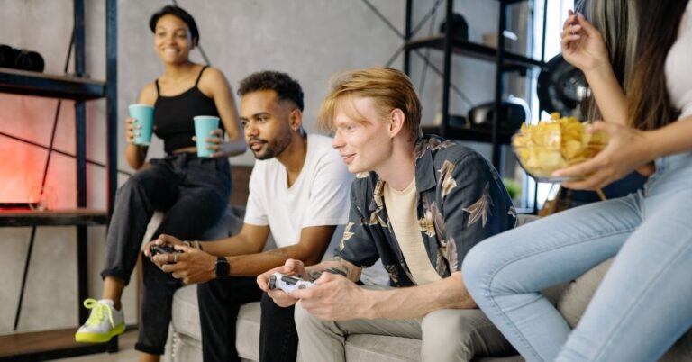 Group of friends playing video games together in a living room, with controllers, drinks, and snacks.
