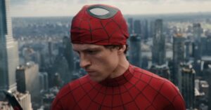 A young man in a red web-patterned superhero suit with his mask rolled up, looking down against a blurred city skyline.